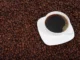 A white coffee cup filled with black coffee, set on a white saucer, surrounded by a large quantity of dark roasted coffee beans.