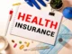 A notebook with "HEALTH INSURANCE" written in bold red and black letters, surrounded by medical items including pills, a thermometer, a face mask, and a pair of blue gloves.