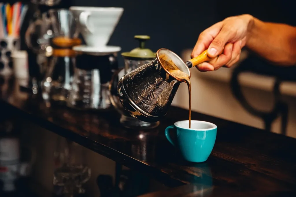 A hand pours dark, rich coffee from an ornate metal cezve (Turkish coffee pot) into a small, vibrant teal cup on a dark wooden counter, with various coffee brewing equipment blurred in the background.
