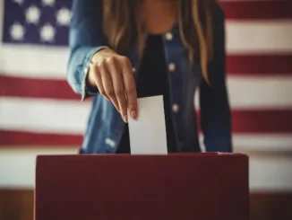 A person's hand casts a white ballot into a red ballot box, with a blurred American flag in the background, symbolizing voting in the U.S. House Election.