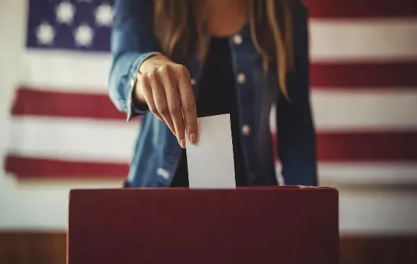 The Power of the Ballot: Participating in Democracy A person's hand casts a white ballot into a red ballot box, with a blurred American flag in the background, symbolizing voting in the U.S. House Election.