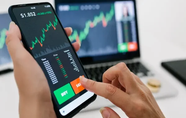 Hands holding a smartphone displaying a cryptocurrency trading chart with "BUY" and "SELL" buttons, against a blurred background of a laptop also showing a crypto chart.