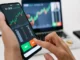 Hands holding a smartphone displaying a cryptocurrency trading chart with "BUY" and "SELL" buttons, against a blurred background of a laptop also showing a crypto chart.
