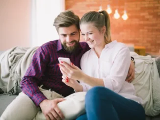 A smiling young couple sitting closely together on a gray couch, looking at a smartphone.