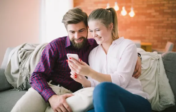 Couple Enjoying Time Together on Couch A smiling young couple sitting closely together on a gray couch, looking at a smartphone.