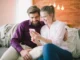 A smiling young couple sitting closely together on a gray couch, looking at a smartphone.
