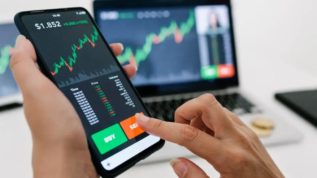 Hands interacting with a cryptocurrency trading app on a smartphone, showing price charts and buy/sell options, with a laptop displaying similar data in the background.