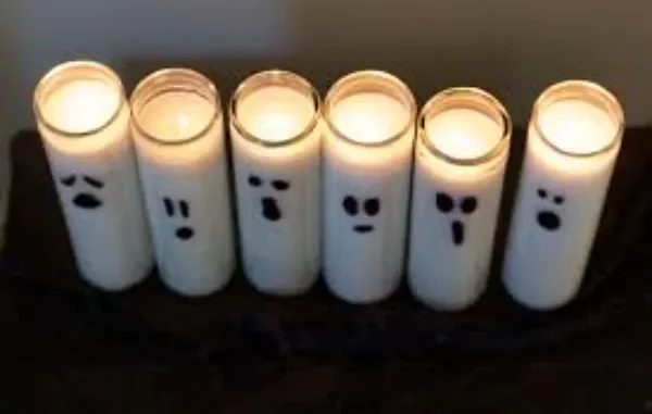 Six white jar candles with simple black marker ghost faces sit lit on a black surface, creating a spooky Halloween decoration.