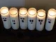 Six white jar candles with simple black marker ghost faces sit lit on a black surface, creating a spooky Halloween decoration.