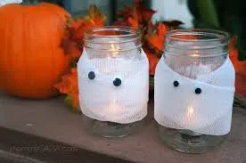 Two glass jars wrapped in white bandages with googly eyes, decorated as mummy candles. They are placed next to a pumpkin and autumn leaves.