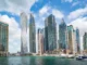 The modern Dubai Marina skyline featuring luxury skyscrapers and yachts docked in the harbor under a bright blue sky with scattered clouds.