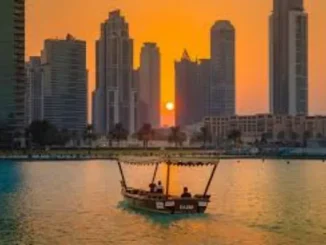 A traditional abra boat cruises on a calm waterway in Dubai at sunset, silhouetted against modern high-rise buildings and a vibrant orange sky.