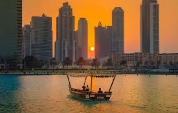 Dubai Sunset: Navigating the True Cost of Living A traditional abra boat cruises on a calm waterway in Dubai at sunset, silhouetted against modern high-rise buildings and a vibrant orange sky.