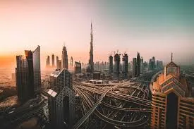 A wide aerial view of the Dubai skyline at sunrise or sunset, showcasing numerous modern skyscrapers, including the Burj Khalifa, and a complex network of highways. The sky is a gradient of warm oranges and soft blues.