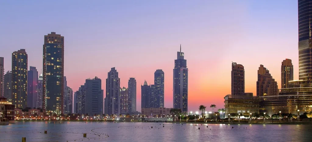 A panoramic view of the Dubai skyline at dusk, with illuminated skyscrapers lining the waterfront under a gradient sky of purple, pink, and orange.
