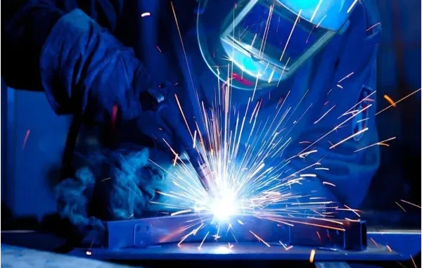 A welder wearing protective gear uses a bright, sparking welding torch on a metal workpiece.