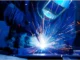 A welder wearing protective gear uses a bright, sparking welding torch on a metal workpiece.