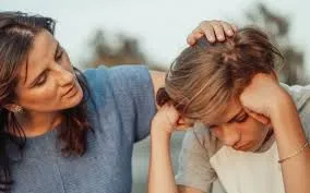 A concerned mother places a comforting hand on the head of her upset teenage child.