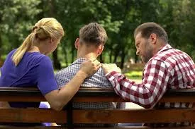 Two parents sit on a park bench with their teenage child, each placing a comforting hand on the teen's shoulders in a show of support.