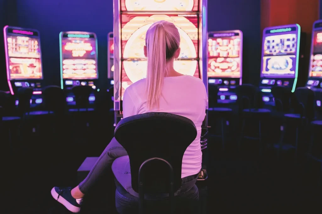 A person sits in front of a brightly lit slot machine in a casino, viewed from behind, highlighting the immersive experience of engaging with slot game features.