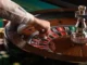 A croupier's hand launching a small white ball into a spinning roulette wheel, while colorful casino chips are visible in the background.