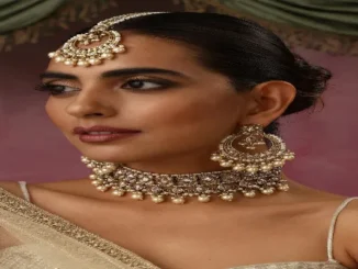 A close-up studio portrait of a woman wearing a stunning traditional Indian bridal jewelry set. She wears a heavily embellished choker necklace adorned with intricate designs and strings of small, creamy white pearls. The choker is complemented by matching large, circular chandelier earrings and a maang tikka, which is a headpiece with a central pendant on the forehead. The jewelry is a rich gold and silver tone with detailed stonework. The woman has her hair styled back and wears subtle makeup with defined eyes. A hint of her sheer, white and gold saree is visible on her shoulder.