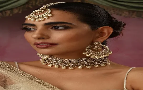 Exquisite Indian Choker and Jewelry Set A close-up studio portrait of a woman wearing a stunning traditional Indian bridal jewelry set. She wears a heavily embellished choker necklace adorned with intricate designs and strings of small, creamy white pearls. The choker is complemented by matching large, circular chandelier earrings and a maang tikka, which is a headpiece with a central pendant on the forehead. The jewelry is a rich gold and silver tone with detailed stonework. The woman has her hair styled back and wears subtle makeup with defined eyes. A hint of her sheer, white and gold saree is visible on her shoulder.