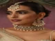 A close-up studio portrait of a woman wearing a stunning traditional Indian bridal jewelry set. She wears a heavily embellished choker necklace adorned with intricate designs and strings of small, creamy white pearls. The choker is complemented by matching large, circular chandelier earrings and a maang tikka, which is a headpiece with a central pendant on the forehead. The jewelry is a rich gold and silver tone with detailed stonework. The woman has her hair styled back and wears subtle makeup with defined eyes. A hint of her sheer, white and gold saree is visible on her shoulder.