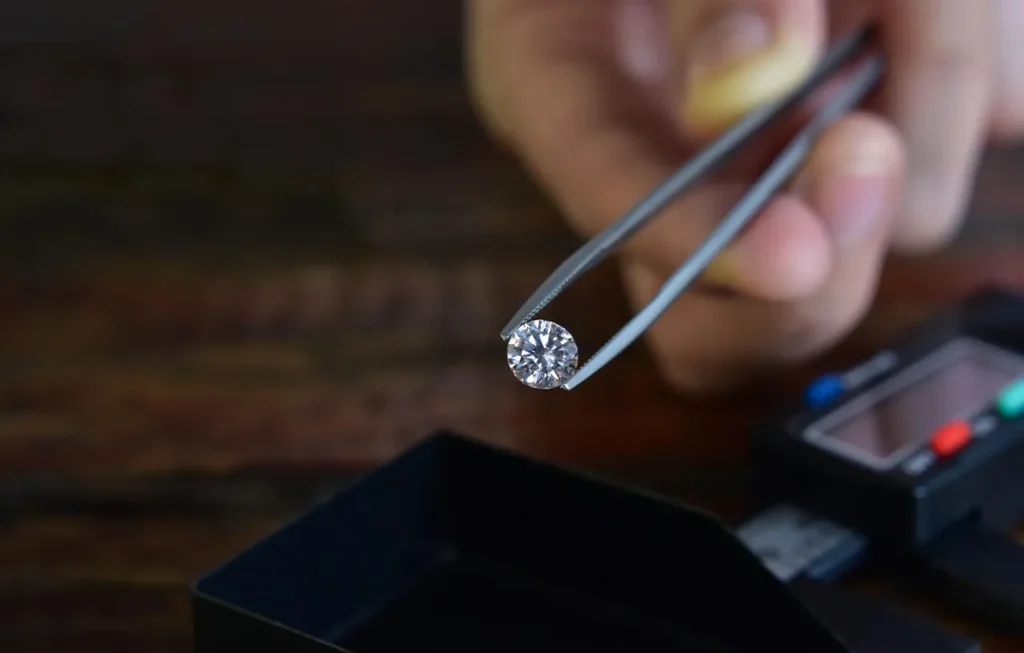 A hand holding a brilliant, round-cut diamond with tweezers, poised above a gemological tool, symbolizing the expert evaluation of engagement ring stones.