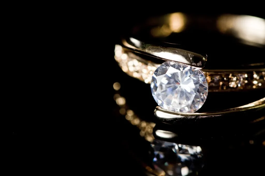 A close-up of a brilliant, round-cut diamond set in a polished gold band, reflecting light against a deep black background, highlighting the stone's radiance.