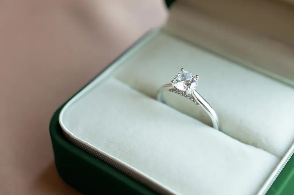 A radiant, round-cut diamond engagement ring with a silver band featuring pavé accents, presented in an open dark green velvet ring box.