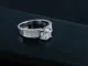 A sparkling silver engagement ring featuring a large, brilliant oval-cut diamond as the center stone, flanked by pavé-set smaller diamonds on a wide band, set against a dark, reflective surface.