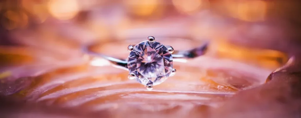 A close-up of a brilliant, round-cut diamond engagement ring with a silver band, resting on a soft, textured pink and gold background, highlighting its sparkle.