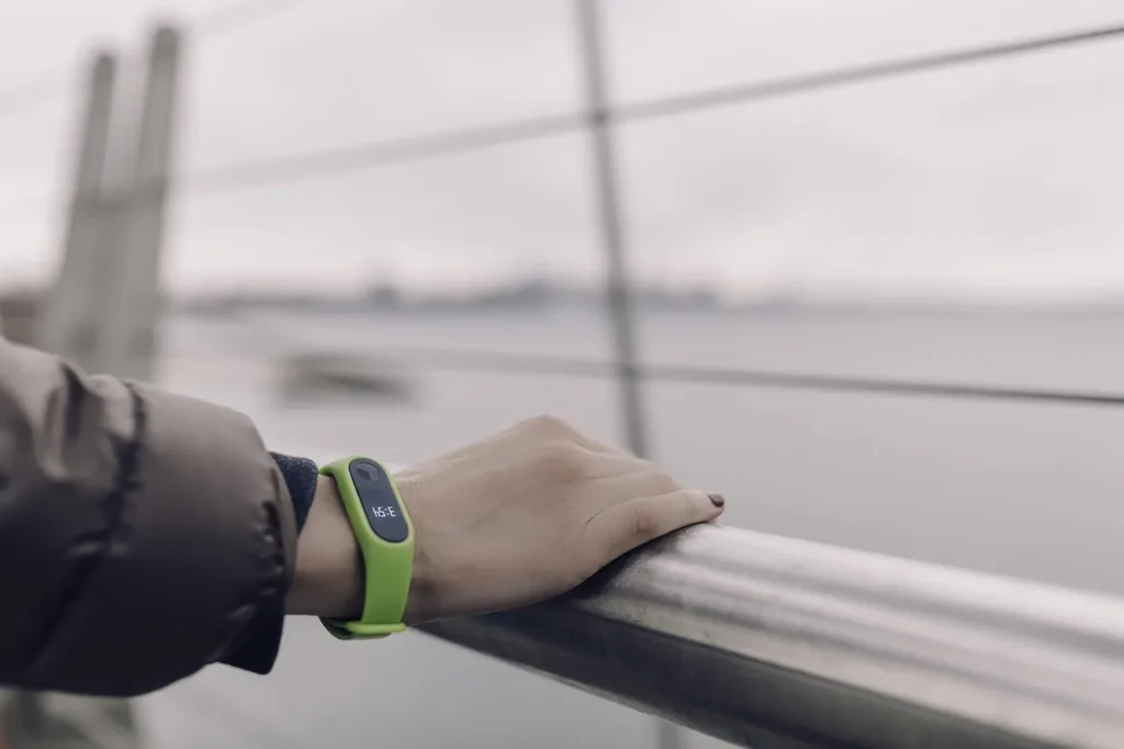 A person's hand, wearing a bright green fitness tracker displaying the time, rests on a metal railing with a blurred outdoor background, representing personal smart automation.