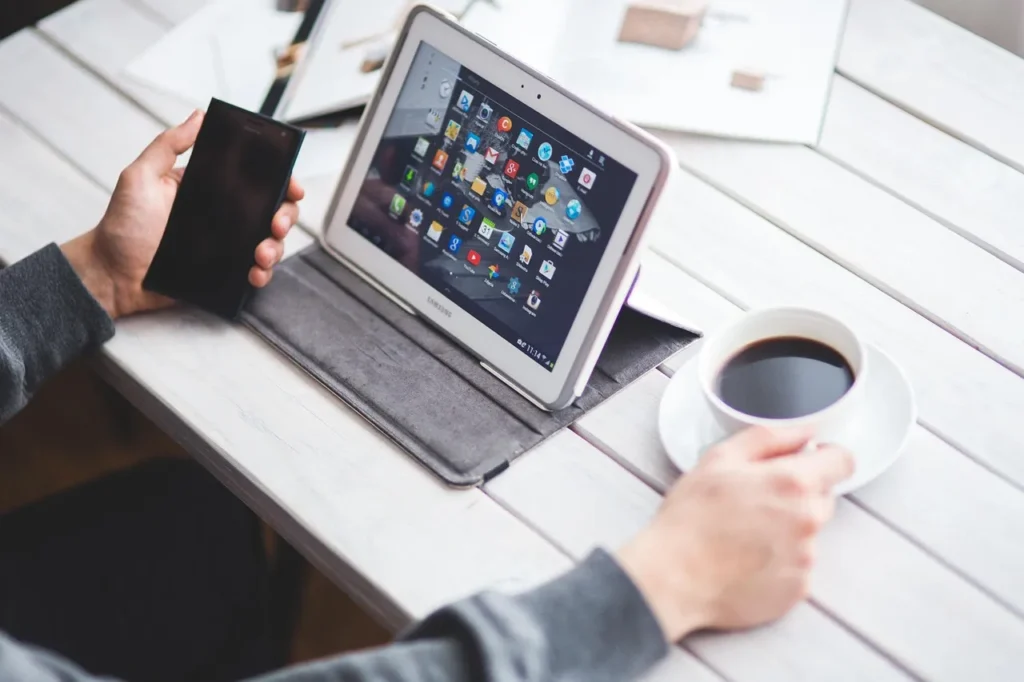 A person's hands holding a black smartphone and a white tablet displaying multiple app icons, with a cup of coffee nearby, illustrating the use and connection of multiple digital devices.