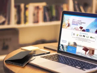 A person's hands are shown at a white desk, holding a black smartphone and a cup of coffee, with a tablet displaying various app icons propped up between them, symbolizing a cross-device professional workflow.