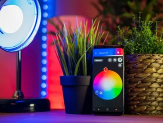A smartphone displaying a smart home app with a color wheel for light control, positioned between two potted plants, with a smart lamp emitting blue light in the background, showcasing home automation.