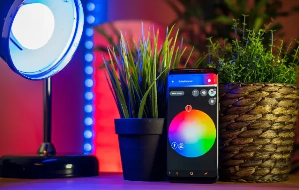 A smartphone displaying a smart home app with a color wheel for light control, positioned between two potted plants, with a smart lamp emitting blue light in the background, showcasing home automation.
