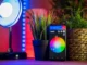 A smartphone displaying a smart home app with a color wheel for light control, positioned between two potted plants, with a smart lamp emitting blue light in the background, showcasing home automation.