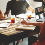 A diverse group of people collaborating around a table with laptops, notebooks, and drinks, representing a strategic product design meeting.