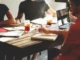 A diverse group of people collaborating around a table with laptops, notebooks, and drinks, representing a strategic product design meeting.