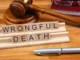 Wooden letter tiles spelling out "WRONGFUL DEATH" are placed in front of a gavel and a red book, symbolizing the legal and judicial process for wrongful death cases.