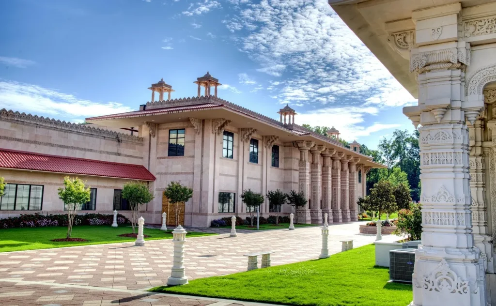 The beige exterior of a large, ornate Hindu temple with multiple carved pillars and towers, surrounded by a manicured green lawn and clear blue sky.