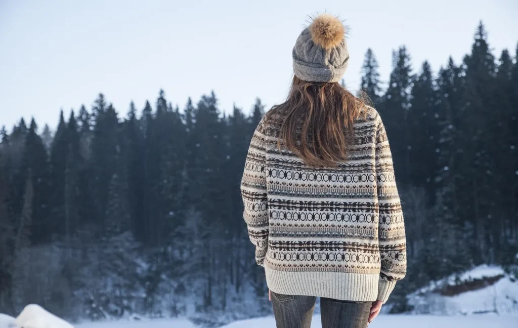 A woman viewed from behind, wearing a thick, patterned Fair Isle or Nordic-style knit sweater and a bobble hat, standing in a snowy landscape with a dense, dark evergreen forest in the background.