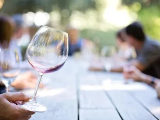 A close-up of a hand holding a stemmed wineglass with a small amount of red wine, tilted slightly, set over a rustic wooden table with people blurred in the background, suggesting an elegant outdoor social gathering.
