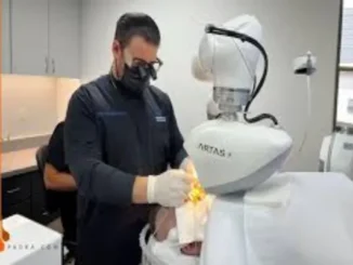 A surgeon wearing a face mask and surgical loupes (magnifying glasses) operates the robotic arm of an ARTAS hair transplant system over a patient lying on a bed.