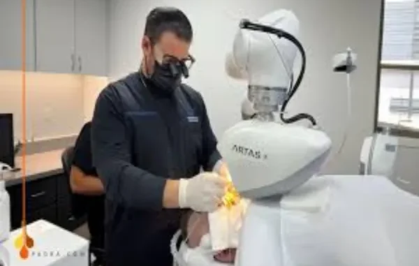Precision and Privacy: The Digital Future of Hair Transplantation A surgeon wearing a face mask and surgical loupes (magnifying glasses) operates the robotic arm of an ARTAS hair transplant system over a patient lying on a bed.