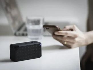 A black portable Bluetooth speaker with a glowing blue light sits on a white desk, while a person's hands in the background hold and interact with a smartphone, symbolizing wireless device connectivity.