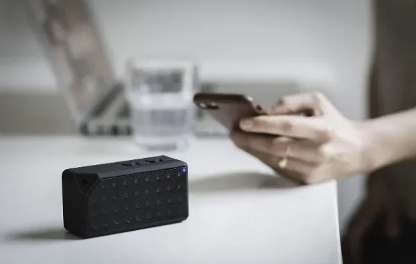 Seamless iPhone and Windows PC Pairing with Start Linking A black portable Bluetooth speaker with a glowing blue light sits on a white desk, while a person's hands in the background hold and interact with a smartphone, symbolizing wireless device connectivity.