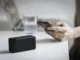 A black portable Bluetooth speaker with a glowing blue light sits on a white desk, while a person's hands in the background hold and interact with a smartphone, symbolizing wireless device connectivity.
